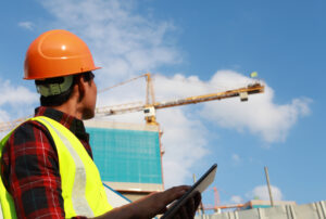 Man looking up at gigantic construction crane and using safety risk prediction software on a tablet.